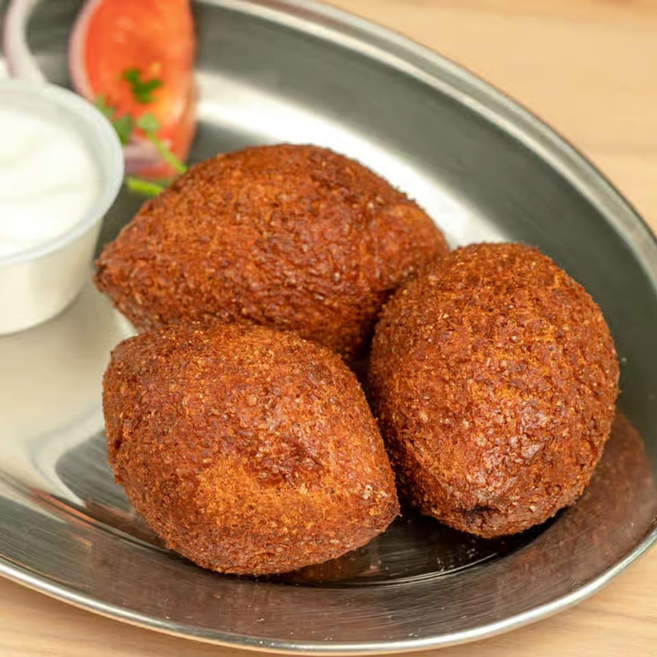 Three golden-brown kibbeh on a silver platter with a side of white sauce and tomato.