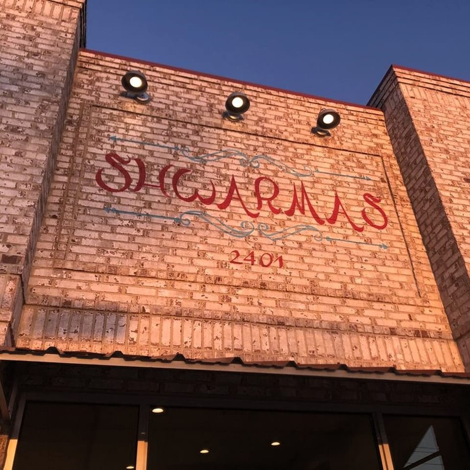 The exterior brick wall of Shwarmas restaurant with its red and blue sign and three spotlights.