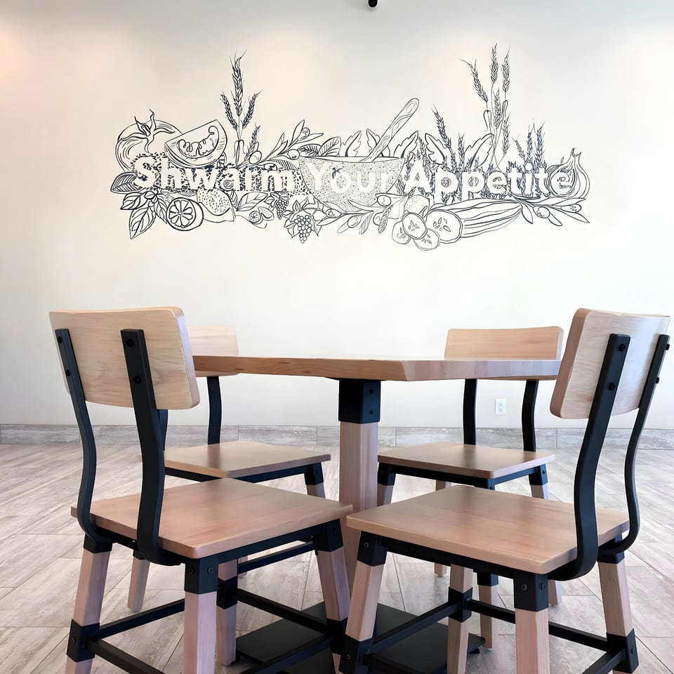 Four wooden chairs and a table in front of a wall mural with Lebanese food illustrations.